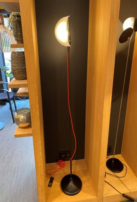 Eine moderne Stehlampe mit halbmondförmigem, weißem Schirm und auffälligem rotem Stromkabel, die in einem hölzernen Ausstellungsregal steht.