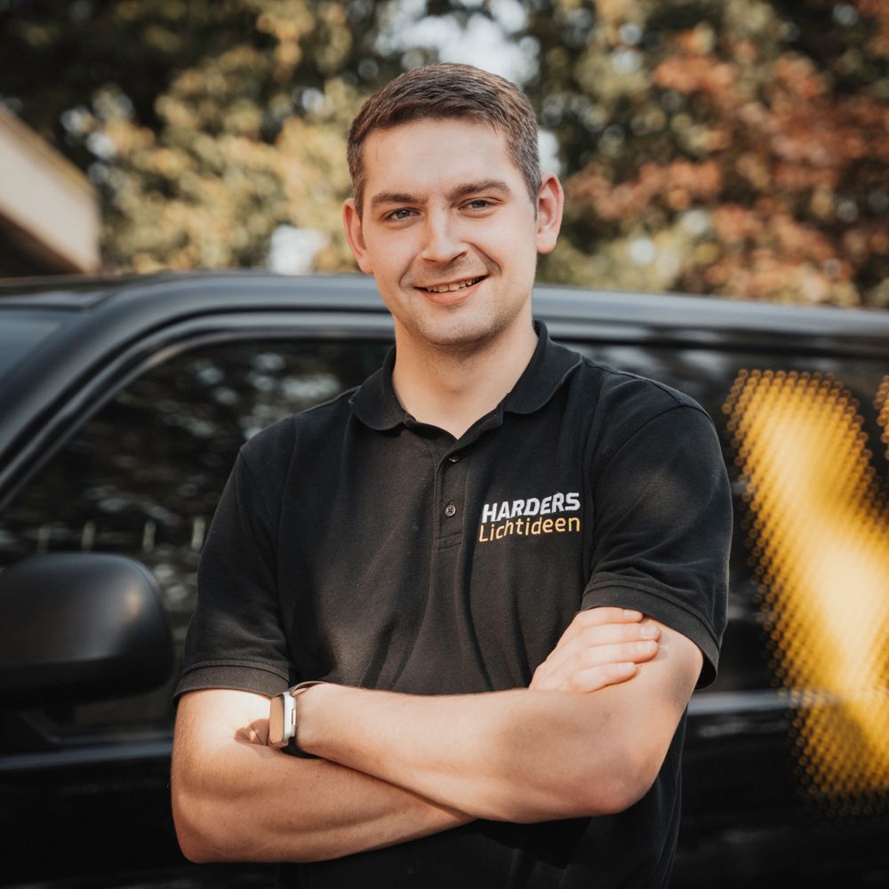Ein Mann in einem schwarzen Hemd steht mit verschränkten Armen vor einem Auto.