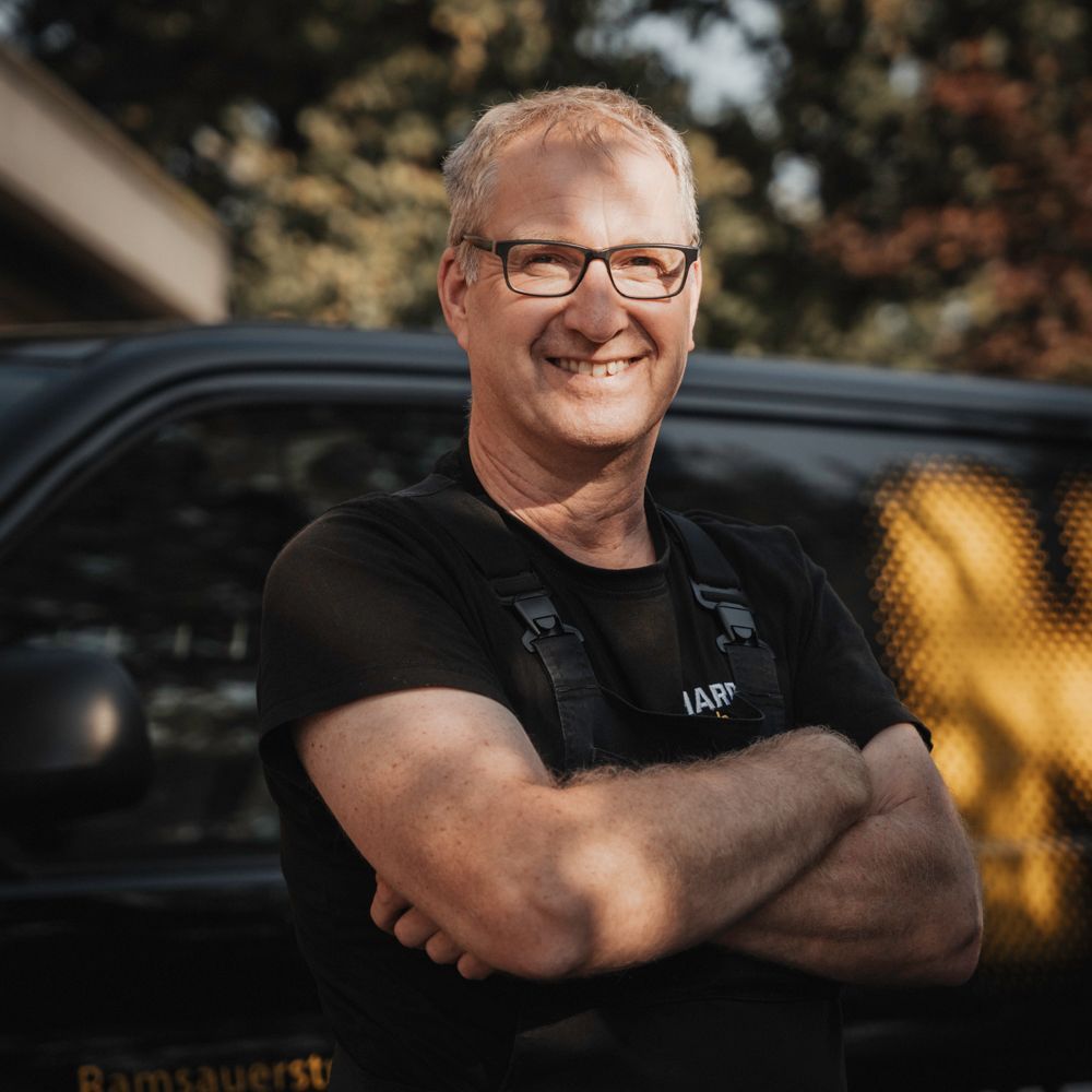 Ein Mann mit Brille und Overall lächelt mit verschränkten Armen