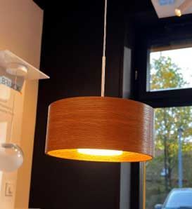 Eine beleuchtete, zylindrische Pendelleuchte in Holzoptik hängt von der Decke. Im Hintergrund ist ein Fenster zu sehen.