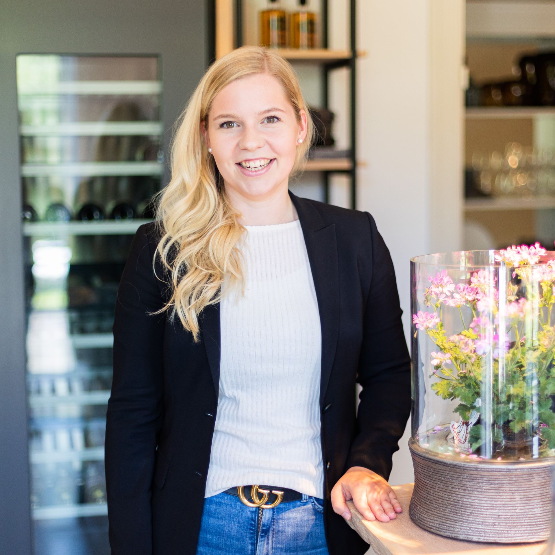 Eine Frau in einer schwarzen Jacke steht neben einer Vase mit Blumen.