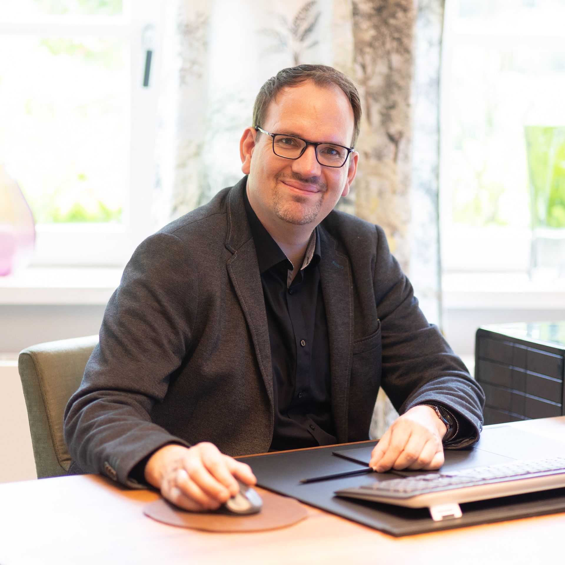 Ein Mann im Anzug und mit Brille sitzt an einem Schreibtisch und benutzt eine Computermaus.