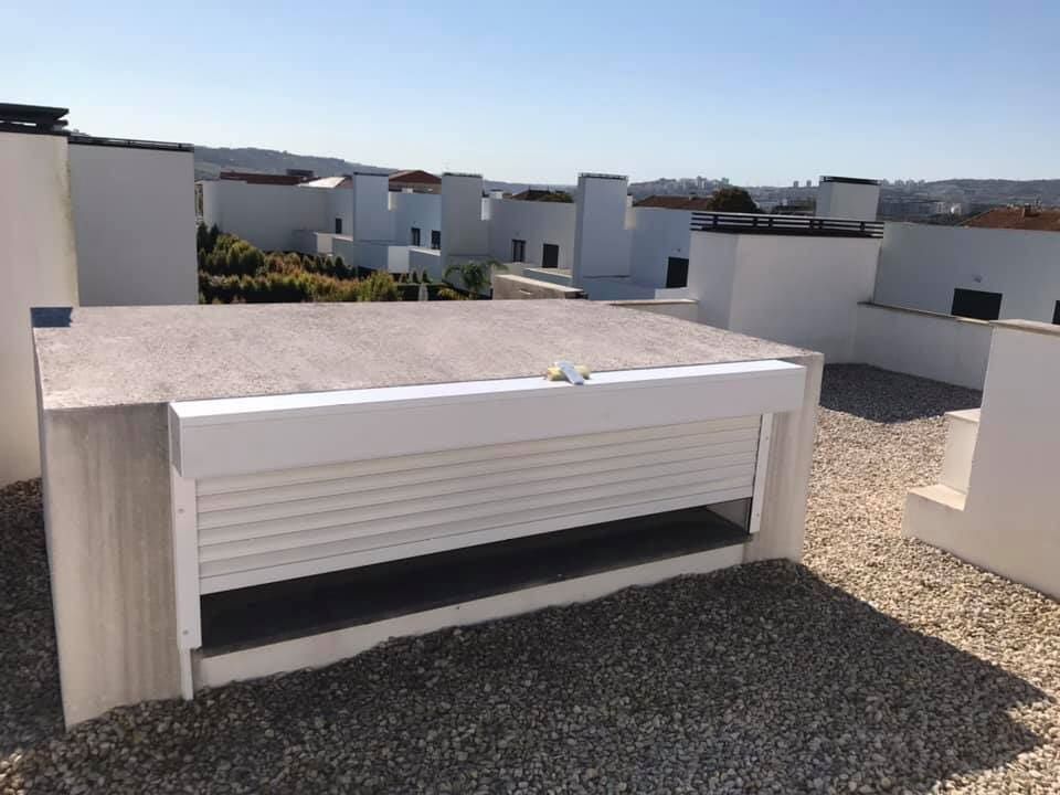 Terraço na cobertura com estrutura branca e persiana aberta; chão de cascalho, dia ensolarado.