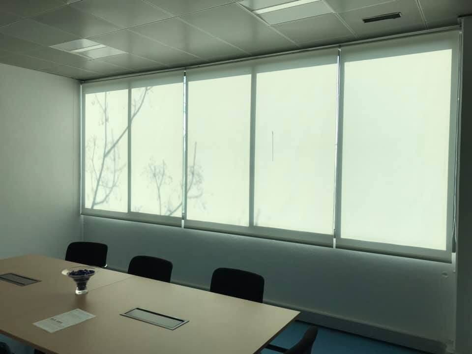 Uma sala de conferências com uma mesa comprida, cadeiras e grandes janelas com persianas brancas fechadas.