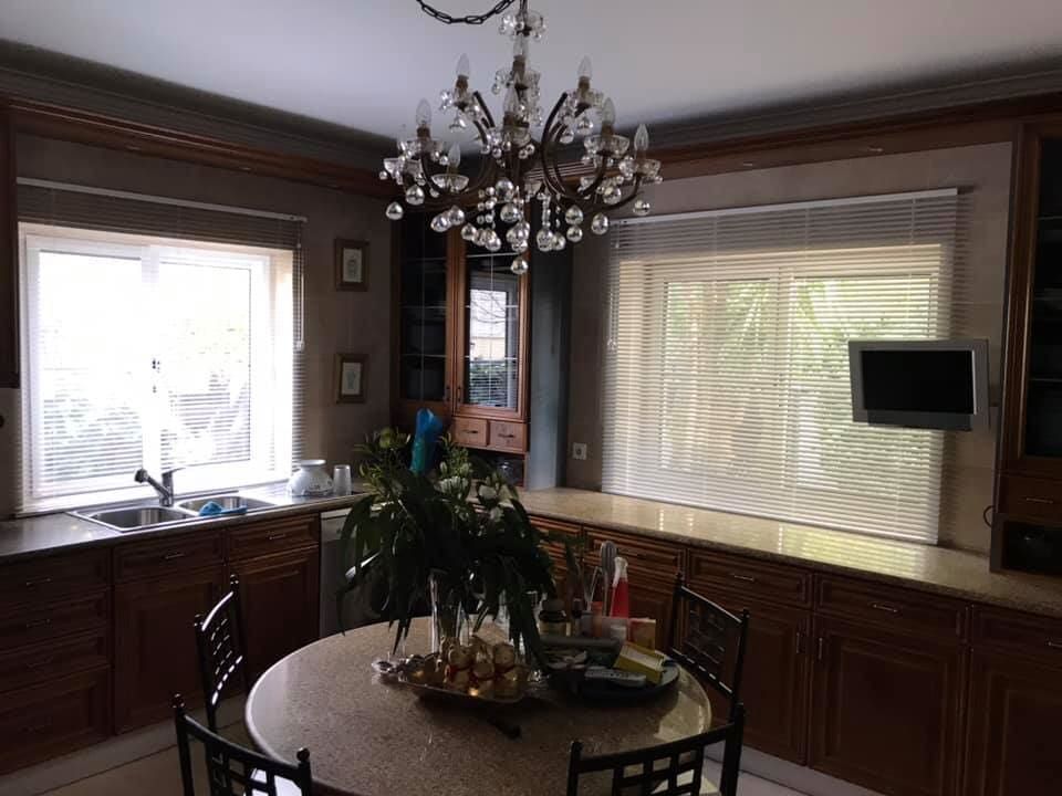 Cozinha com armários de madeira escura, mesa redonda e lustre. Janelas com persianas claras.