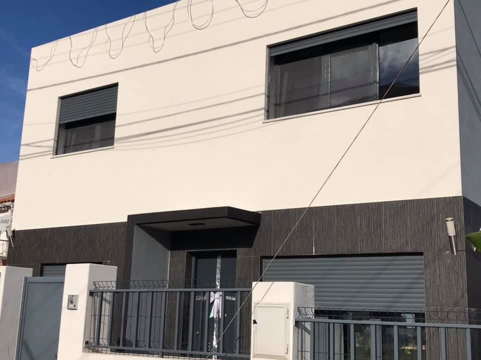 Prédio branco de dois andares com detalhes em cinza, venezianas nas janelas e porta de garagem basculante fechada.