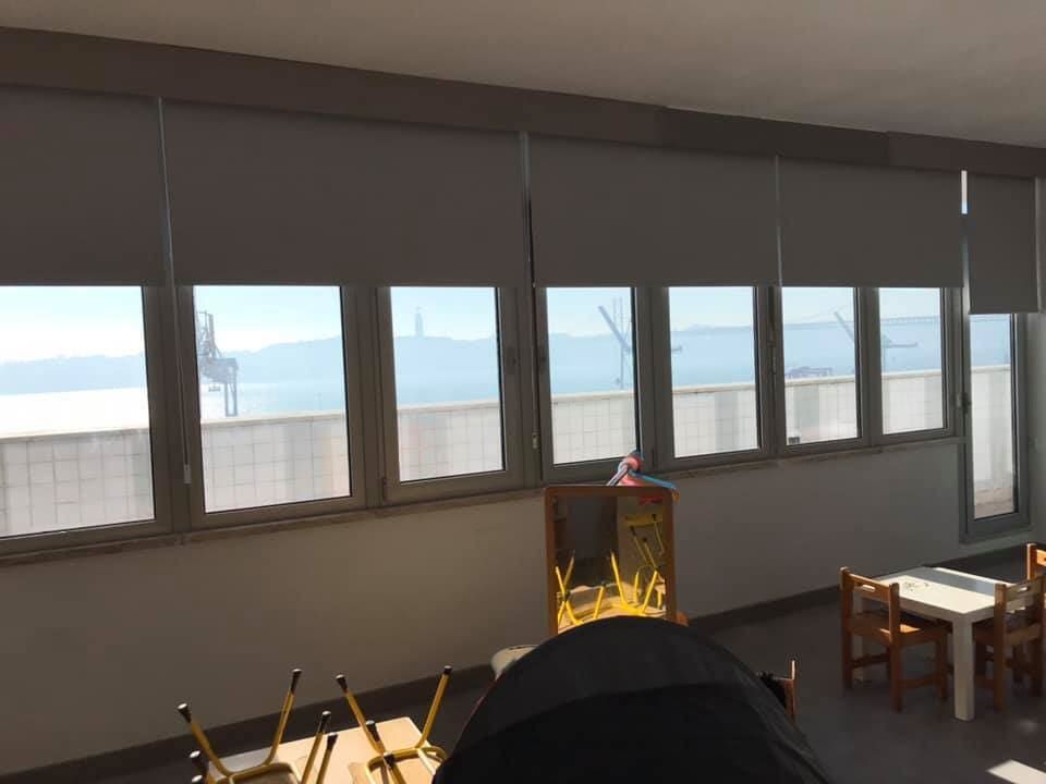 Sala de aula com persianas cinzentas cobrindo parcialmente as janelas com vista para a água. Mobiliário infantil visível.