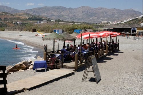 Una playa con mesas y sombrillas y montañas al fondo