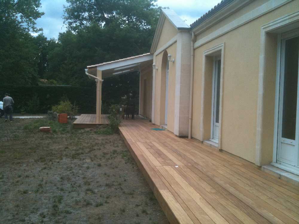 Terrasse en bois longeant une maison beige, à côté d'une cour boueuse et d'arbres.