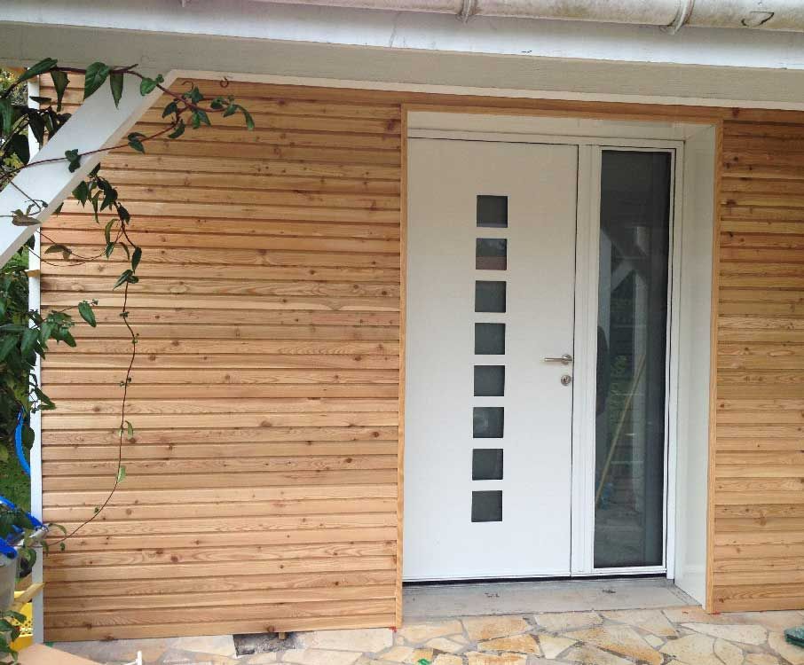 Entrée de maison moderne avec porte blanche et panneau de verre vertical, encadrée par un bardage horizontal en bois.