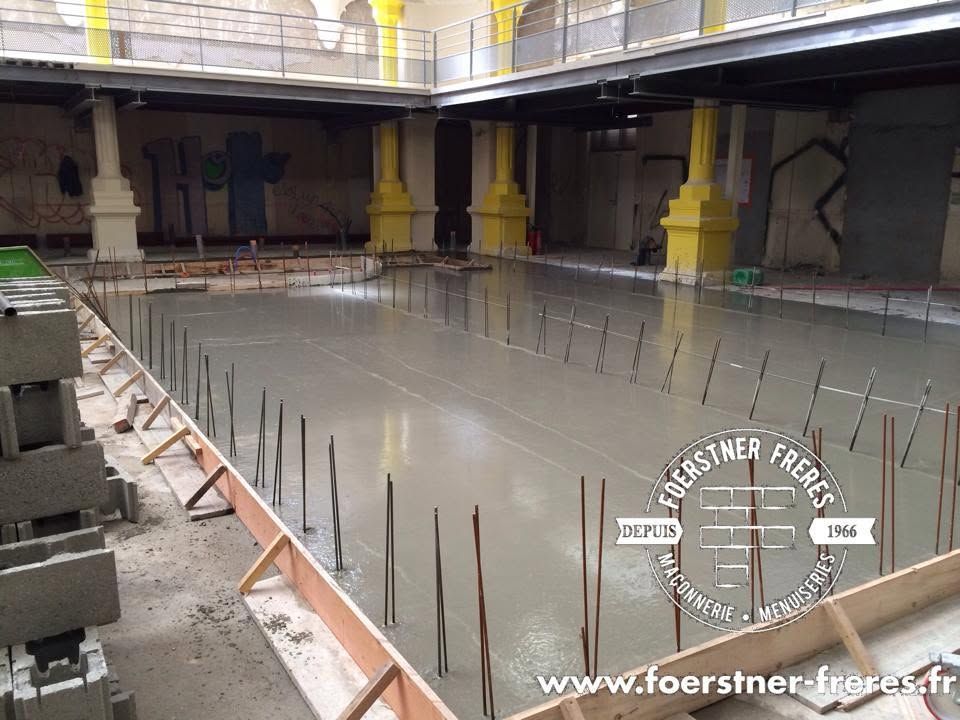 Grande piscine intérieure en construction avec sol en béton, armatures apparentes et colonnes de soutien jaunes.