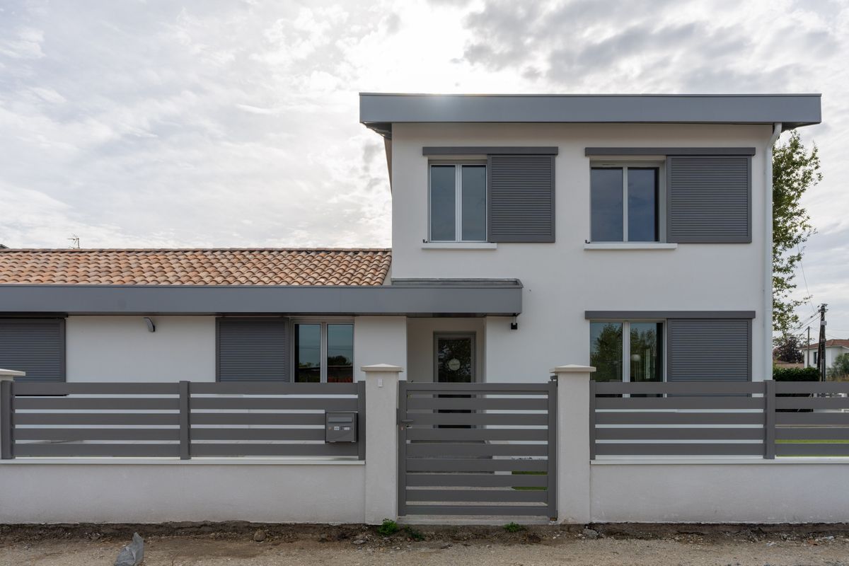 Maison moderne grise à deux étages avec toit de tuiles, clôture à l'avant et entrée fermée par un portail.