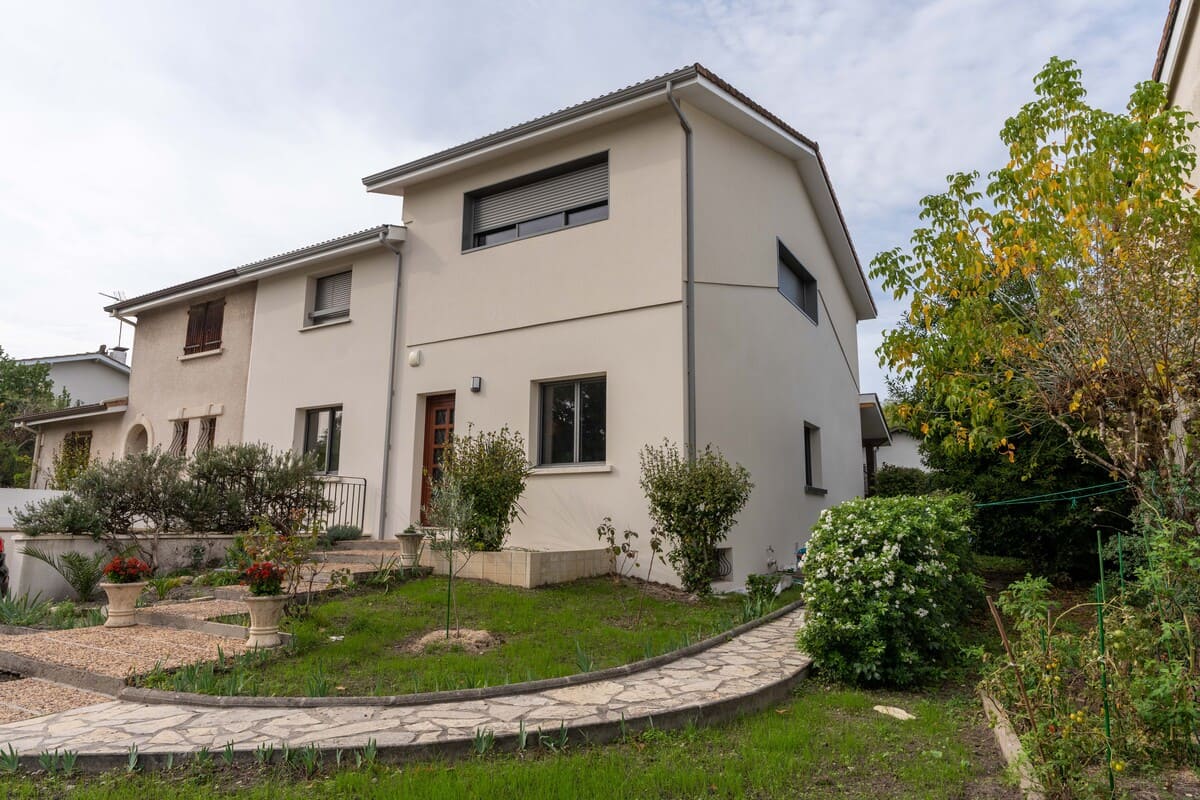 Maison beige à deux étages avec jardin, chemin de pierre et arbres sous un ciel nuageux