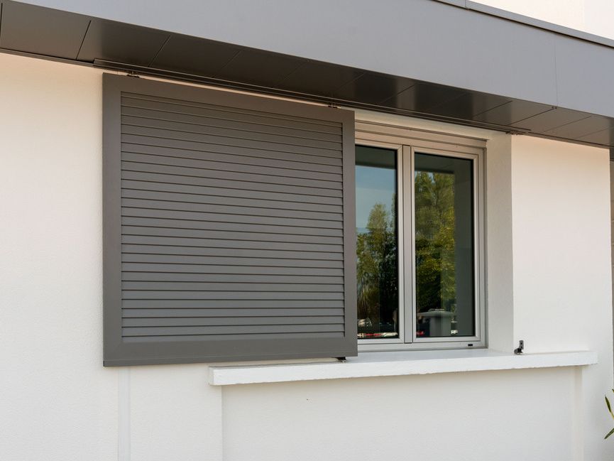 Extérieur d'une maison blanche moderne avec une fenêtre et un volet gris partiellement fermés.