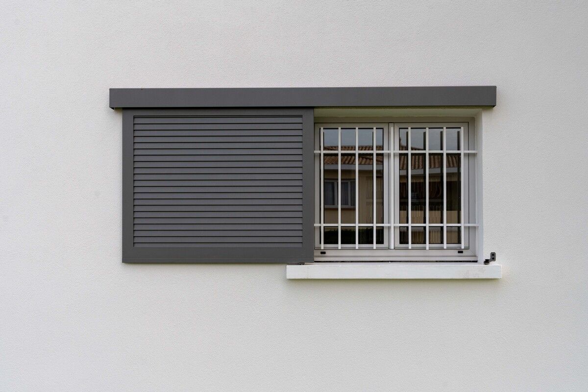 Volet gris fermé à côté d'une fenêtre à barreaux sur un mur blanc