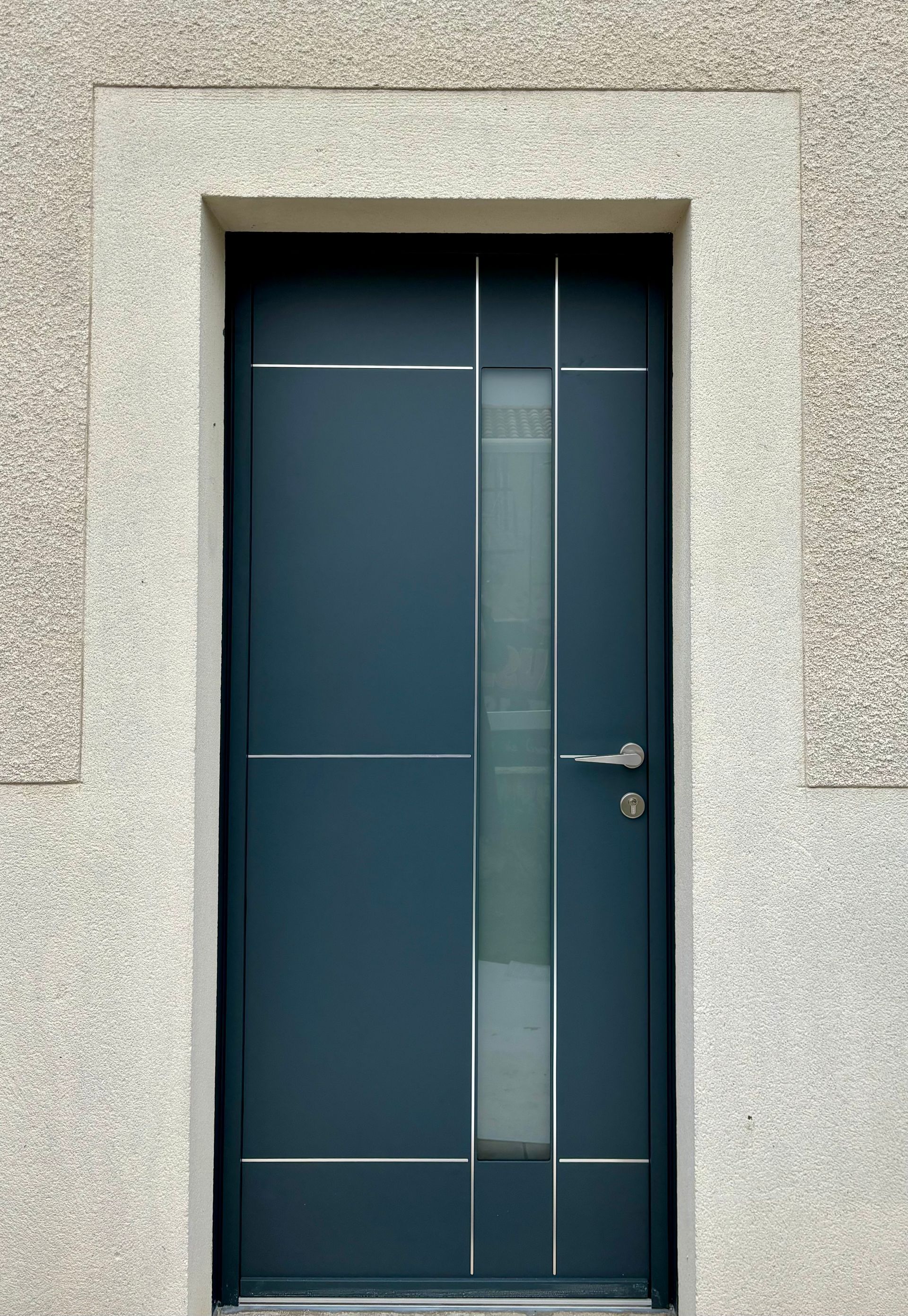 Porte moderne bleu foncé encastrée dans un mur en pierre mouchetée, avec une poignée argentée et un panneau vertical en verre dépoli.