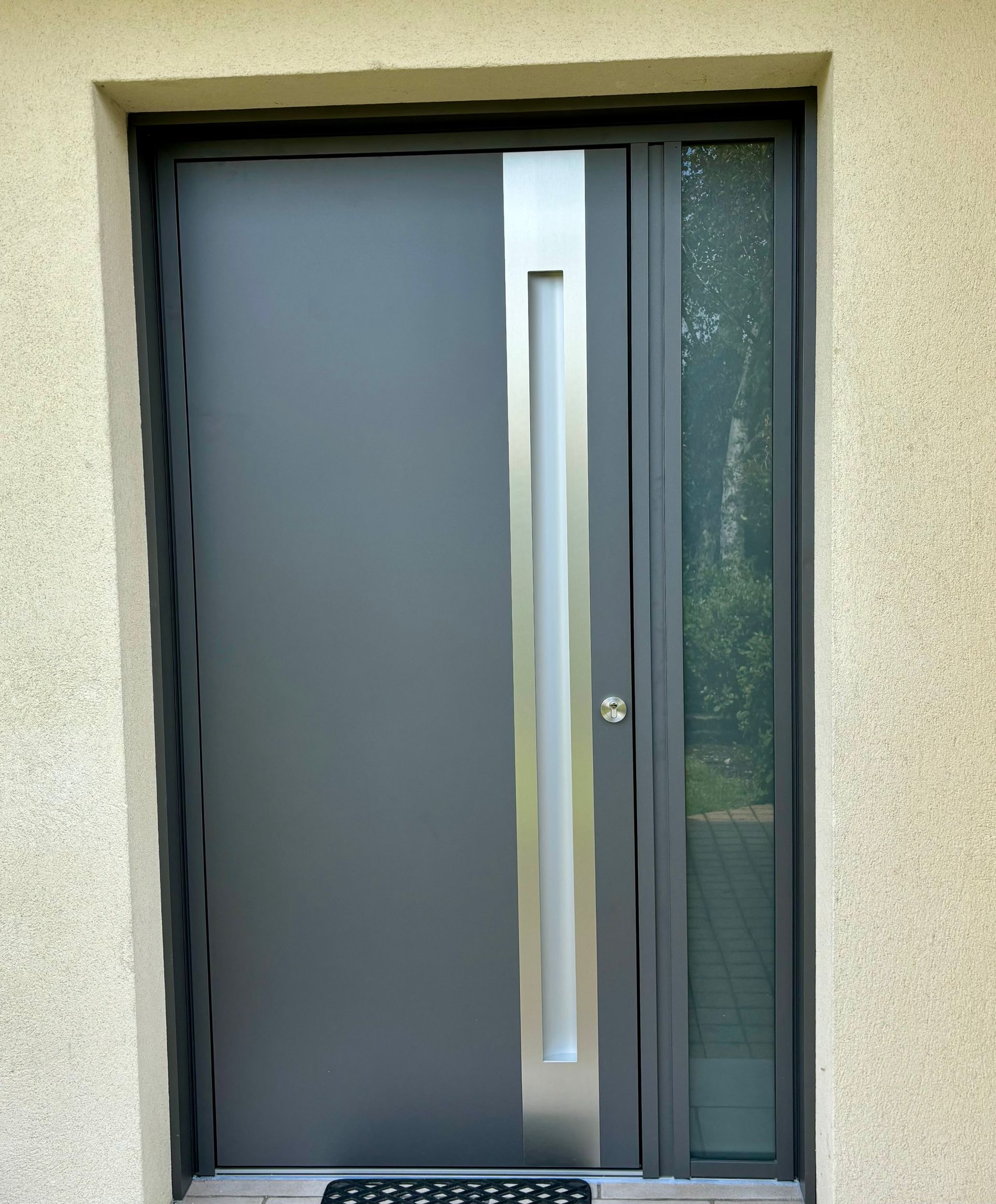 Porte d'entrée grise moderne avec une longue poignée verticale à côté d'un panneau de verre étroit.