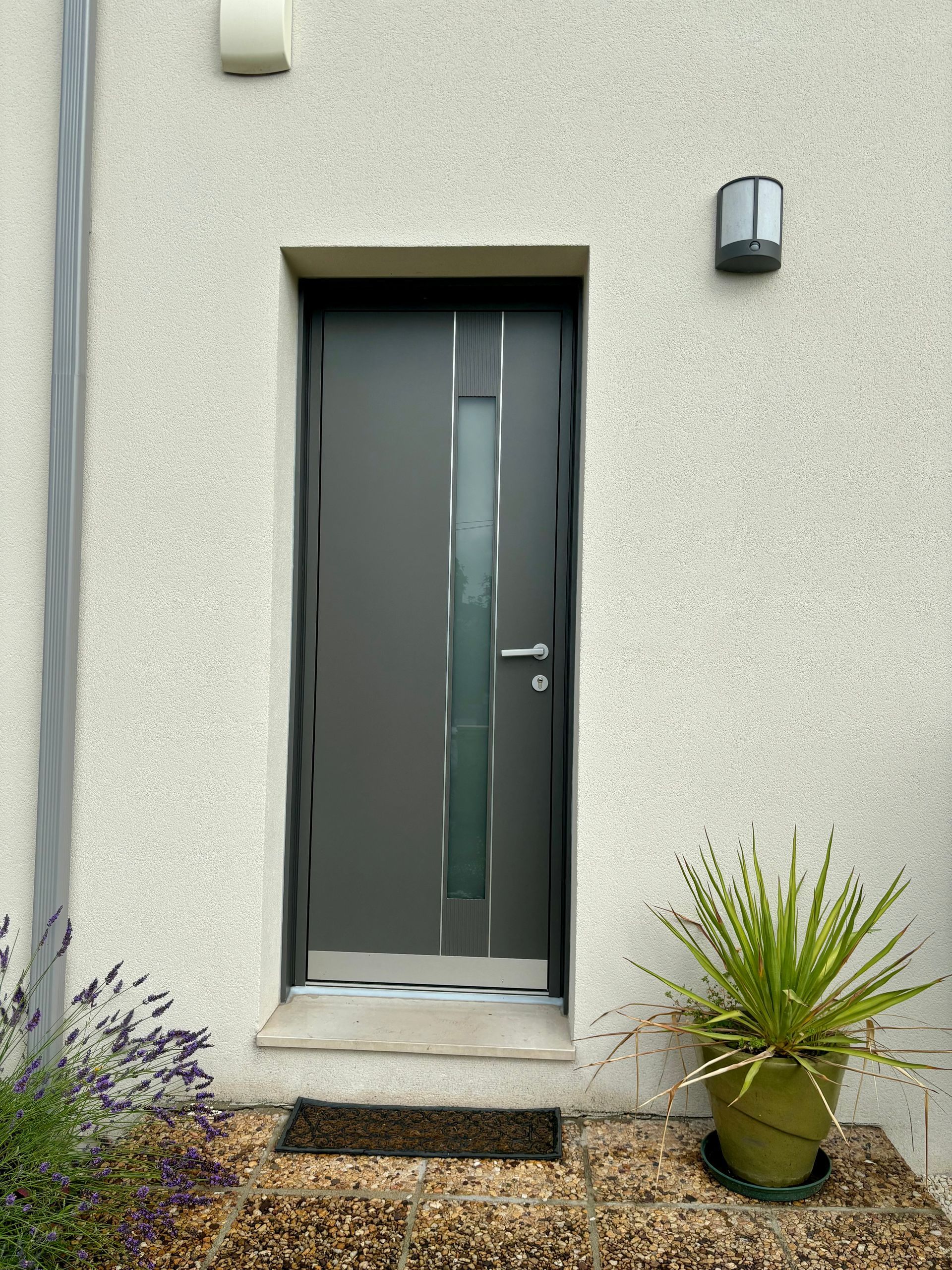 Porte d'entrée grise avec un panneau de verre étroit, flanquée d'appliques murales et de plantes en pot à l'entrée d'une maison