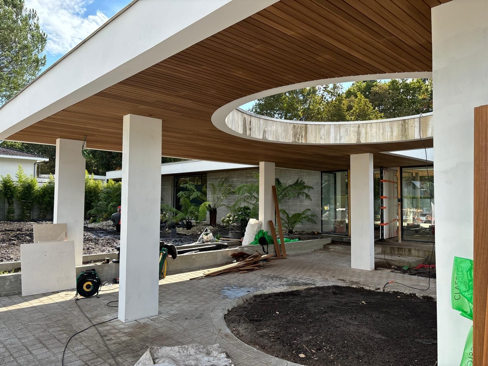 Entrée d'un bâtiment moderne avec auvent en bois incurvé, colonnes blanches et aménagement paysager inachevé.