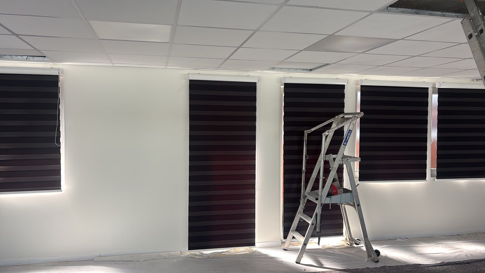 Pièce vide aux murs blancs, aux stores sombres et à un escabeau posé sur un sol clair