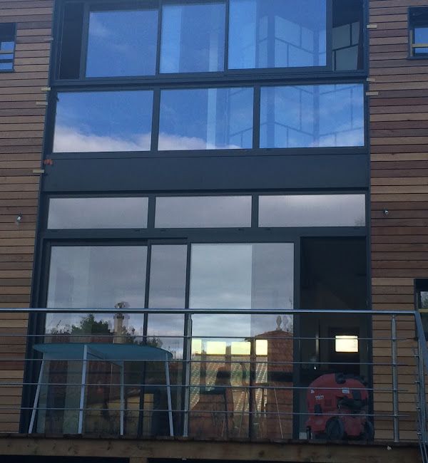 Immeuble d'appartements moderne avec de grandes fenêtres, un balcon et un bardage en bois se reflétant dans une façade vitrée.
