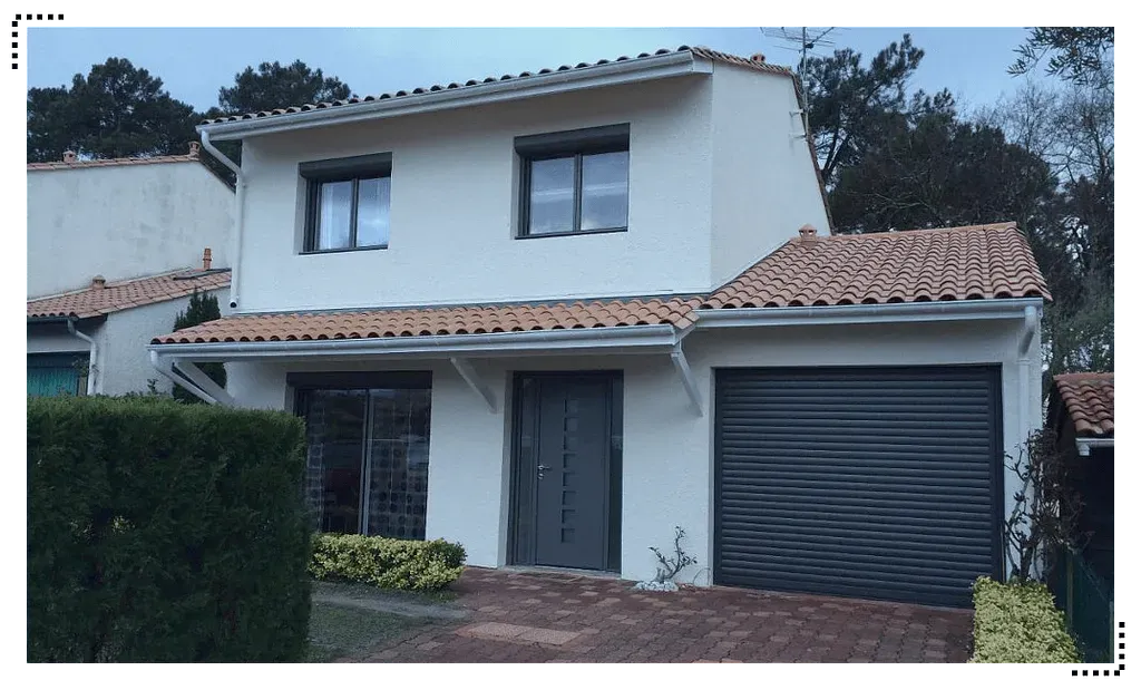 Maison blanche à deux étages avec toit en tuiles de terre cuite et porte de garage grise fermée, entourée de verdure.
