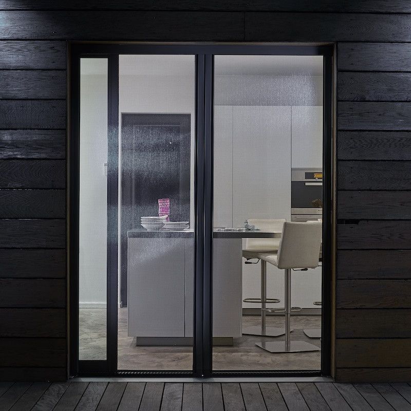 Cuisine moderne vue de nuit à travers une porte vitrée, avec des meubles blancs et un tabouret beige