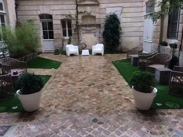 Cour intérieure avec terrasse pavée, chaises blanches et plantes en pot adossée à une façade en pierre