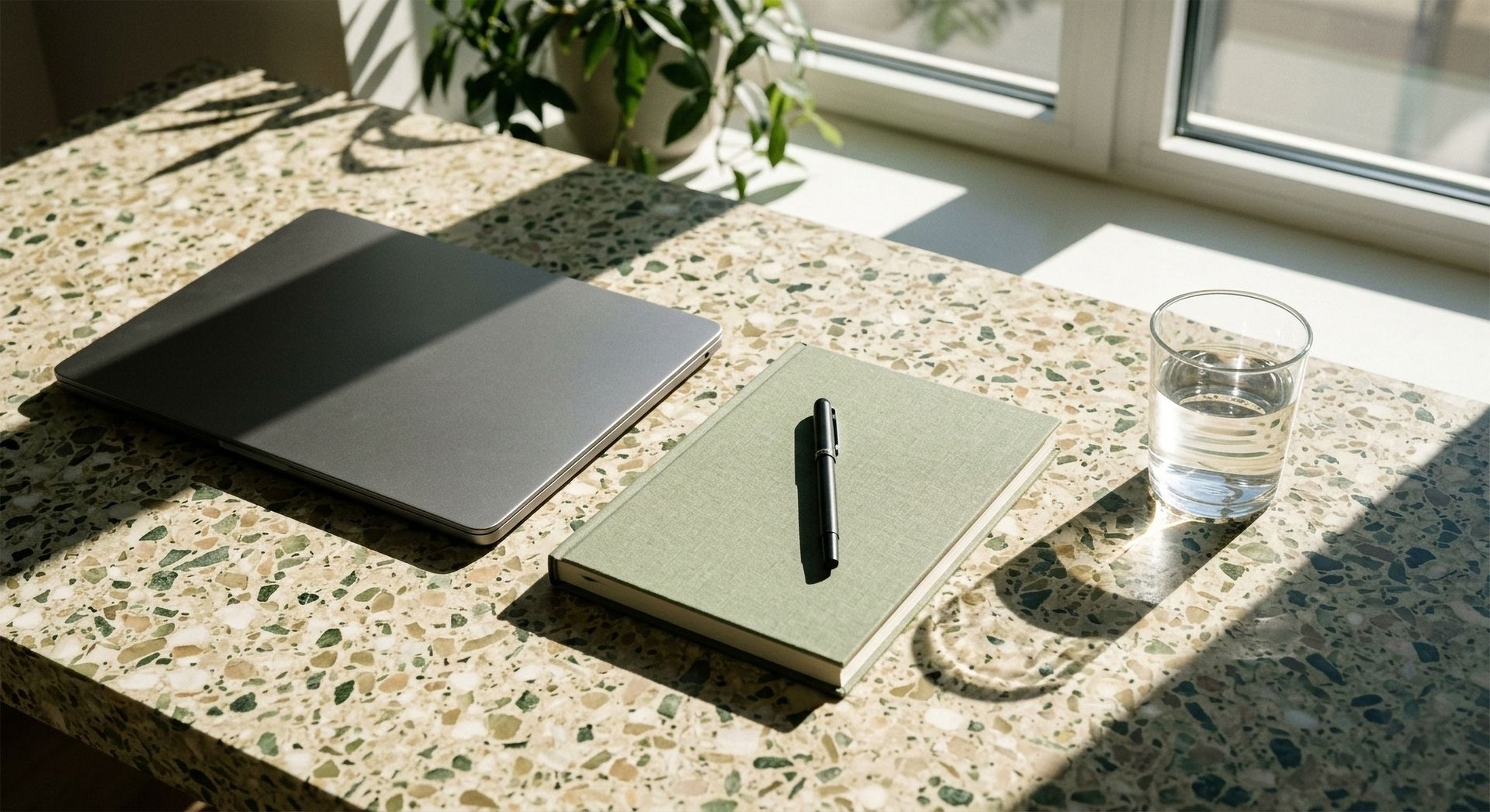 Un carnet et un stylo posés sur un bureau ensoleillé, à côté d'un verre d'eau et d'un ordinateur portable fermé.