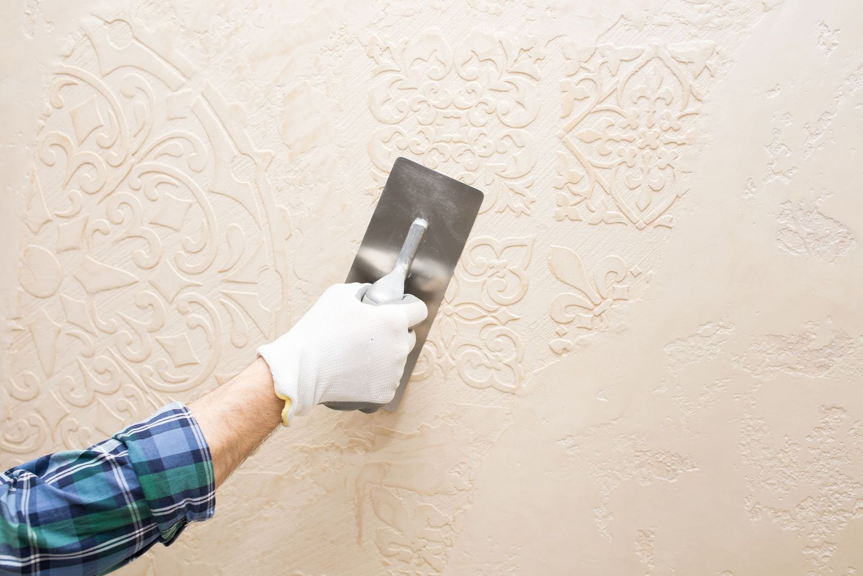 Main gantée lissant un mur beige texturé avec une truelle à mastic en métal.