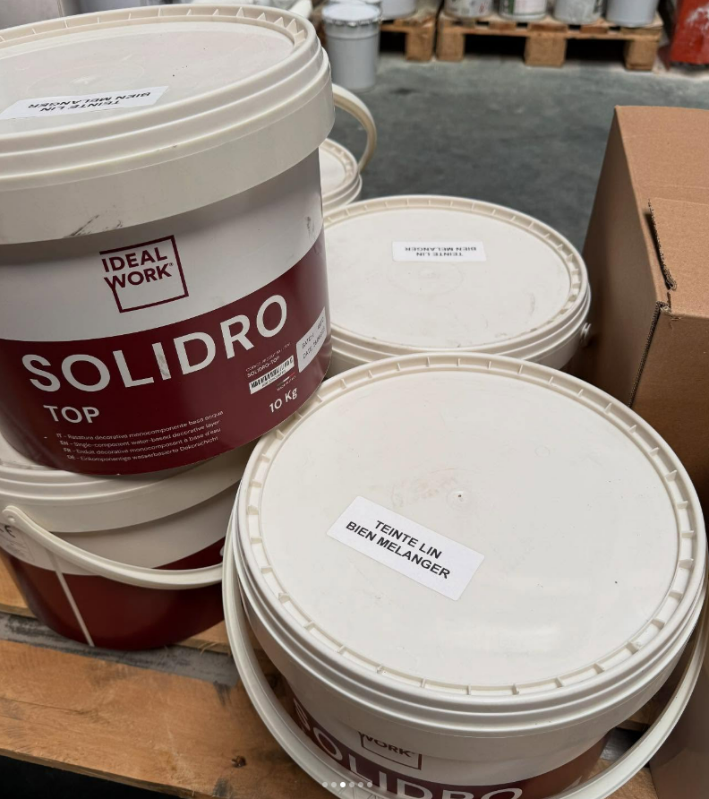 Des seaux de peinture blanche étiquetés SOLIDRO TOP empilés sur une palette en bois dans un entrepôt.