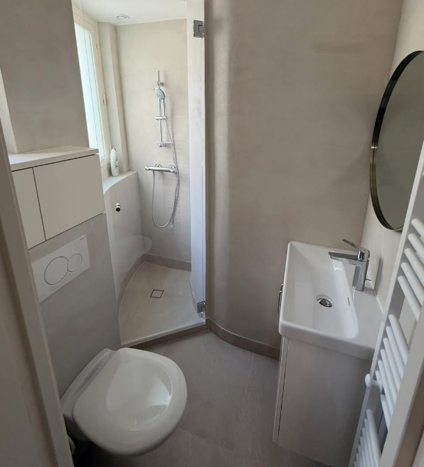 Salle de bains étroite avec toilettes, lavabo et douche dans des tons neutres.