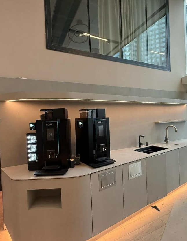 Coin cuisine de bureau avec deux machines à café noires, un évier et des placards en bois clair sous une fenêtre en mezzanine.