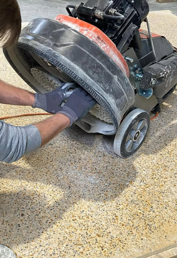Une personne utilise une polisseuse électrique sur un sol moucheté, des mains nettoient des rouleaux sous la machine.