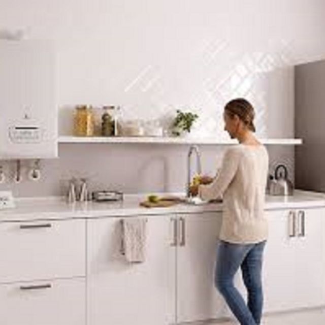 Mujer lavando platos en una cocina blanca y luminosa con un estante lleno de frascos. Caldera en la pared.