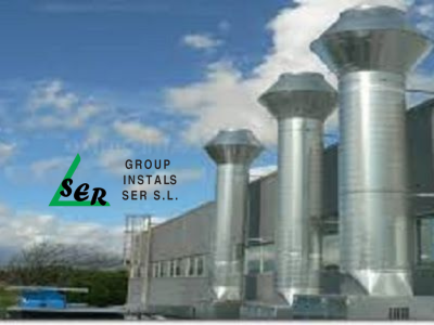 Conductos de escape industriales plateados en el tejado de un edificio, contra un cielo azul con nubes.