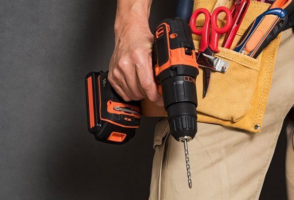 Persona que sostiene un taladro eléctrico naranja y negro y lleva un cinturón de herramientas con tijeras y otras herramientas.