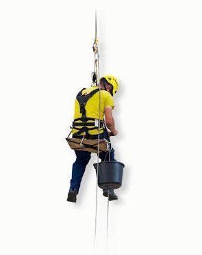 Trabajador con camisa amarilla y equipo de seguridad suspendido de cuerdas con un balde.