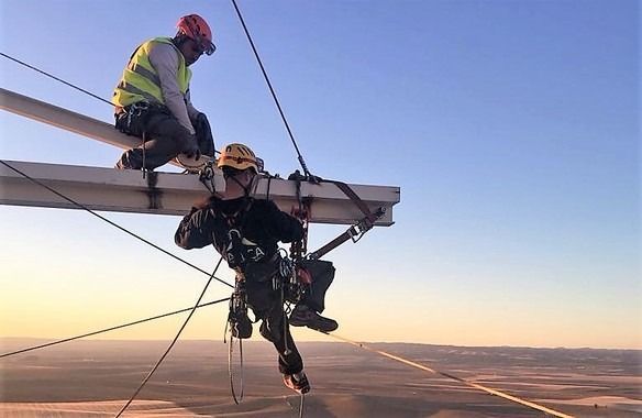 Dos trabajadores en una estructura alta. Uno sentado, el otro suspendido con equipo de seguridad, contra un cielo azul.