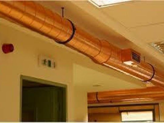 Conductos de aire acondicionado color cobre corren a lo largo del techo, sujetos por correas negras. Se ven una puerta y una luz de emergencia.