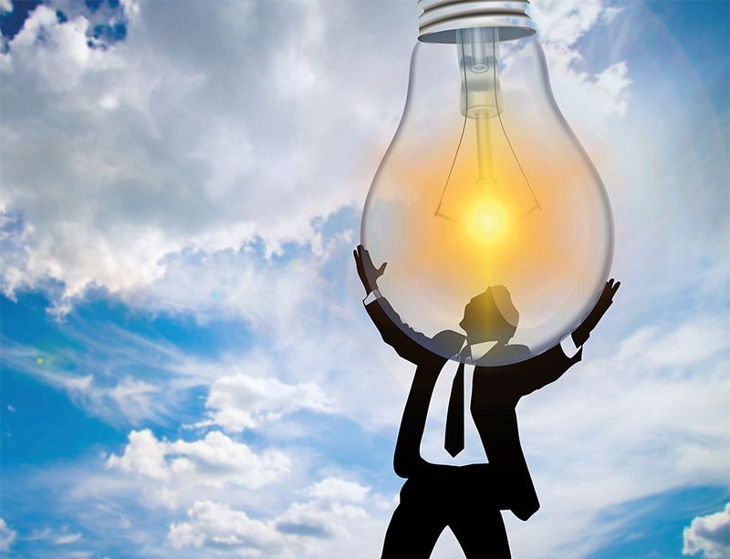 Silueta de una persona sosteniendo una bombilla grande y brillante contra un cielo nublado.