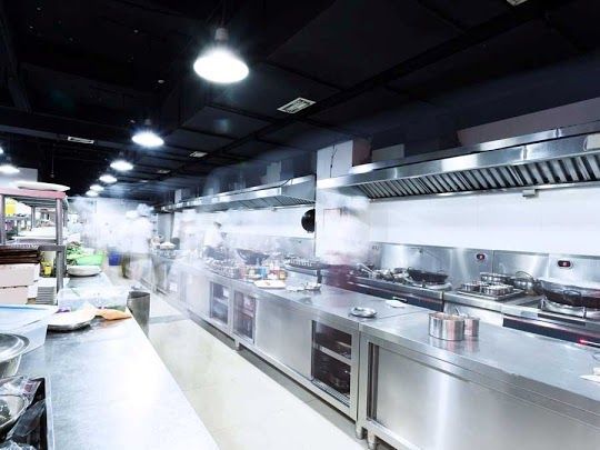 Cocina comercial con encimeras, hornillas y campanas extractoras de acero inoxidable; vapor visible.