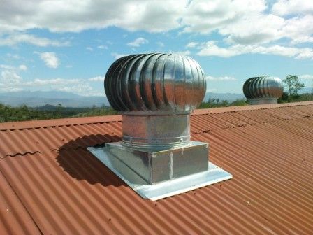 Dos respiraderos de turbina plateados sobre un techo rojo corrugado contra un cielo azul, con una segunda turbina en el fondo.