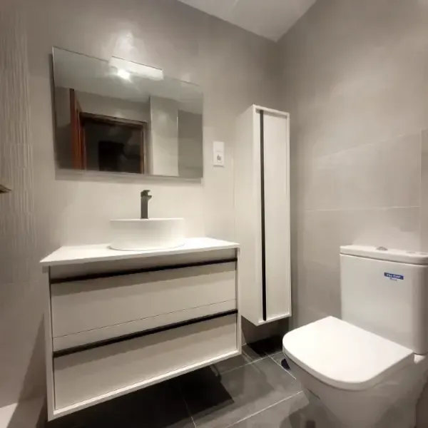 Baño moderno con tocador blanco, lavabo, espejo, mueble alto e inodoro; paredes y suelo grises.