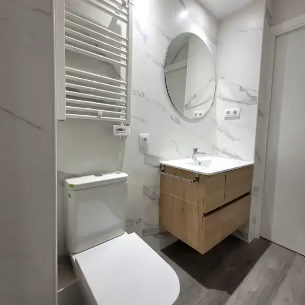 Baño moderno con pared efecto mármol, tocador flotante de madera, espejo ovalado, calentador de toallas e inodoro.