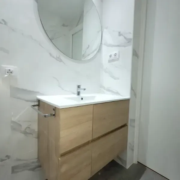 Baño con tocador de madera, encimera blanca y gran espejo redondo sobre revestimiento efecto mármol.