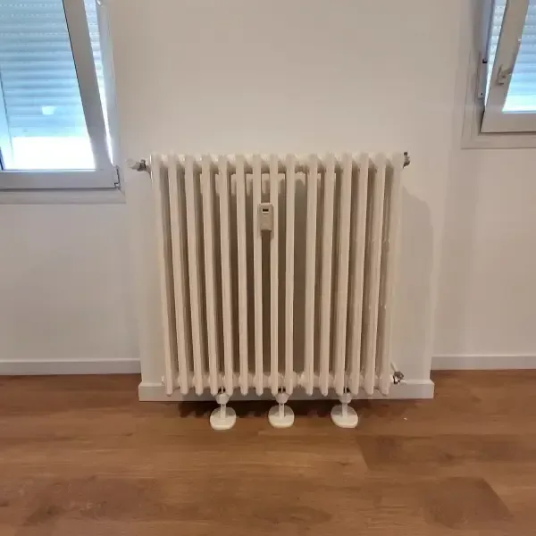 Radiador blanco en una pared entre dos ventanas, sobre un suelo de madera.