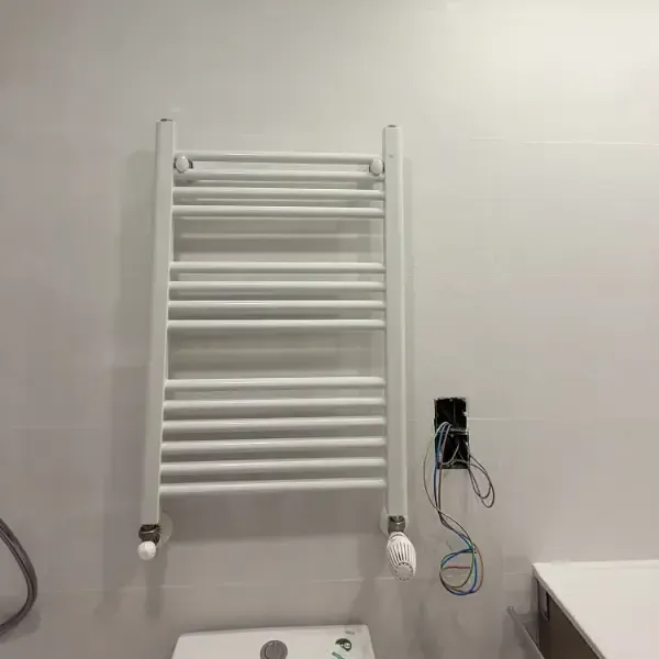Calentador de toallas blanco sobre una pared de azulejos blancos cerca de una caja eléctrica expuesta.