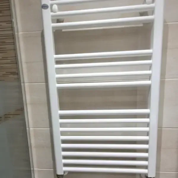 Calentador de toallas blanco montado en una pared de baño con azulejos de color beige.