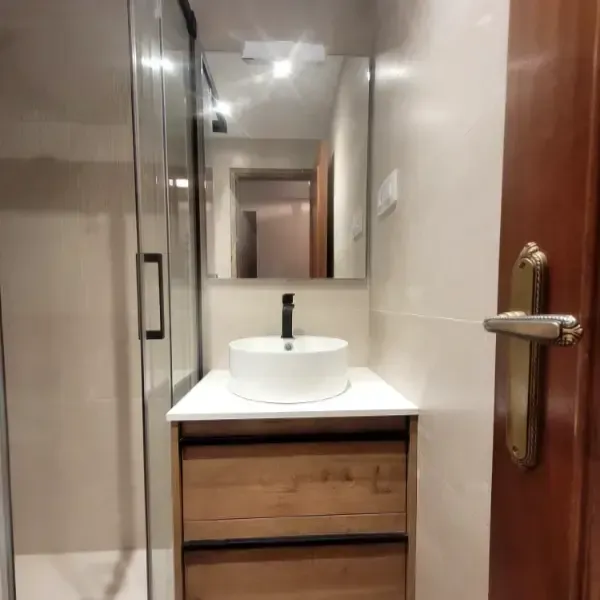 Baño con tocador de madera, lavabo blanco y ducha. Un espejo cuelga sobre el lavabo.